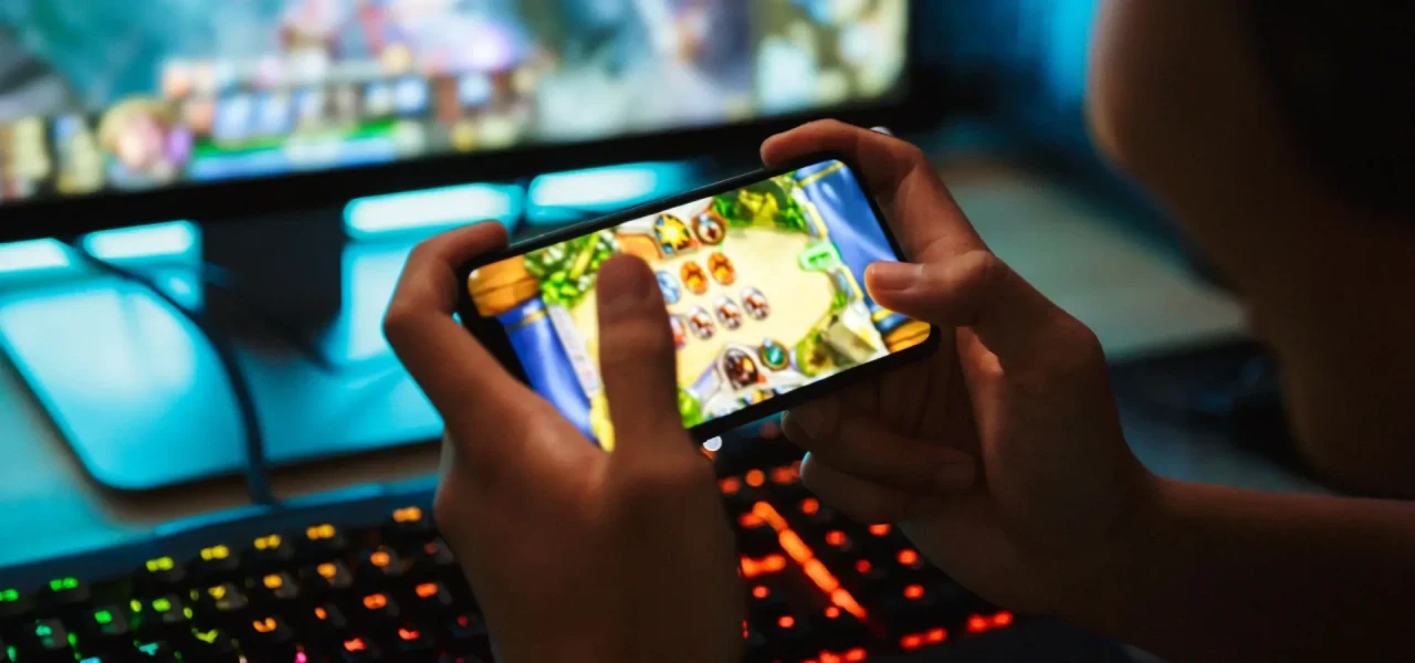 gameverslaving-op-telefoon-en-computer Iemand met een gameverslaving speelt een spelletje op zowel smartphone als computer.