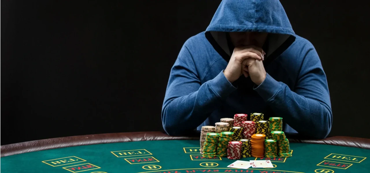 man-met-pokerverslaving Man in blauw vest met pokerverslaving zit achter een groene pokertafel met fiches voor zich.