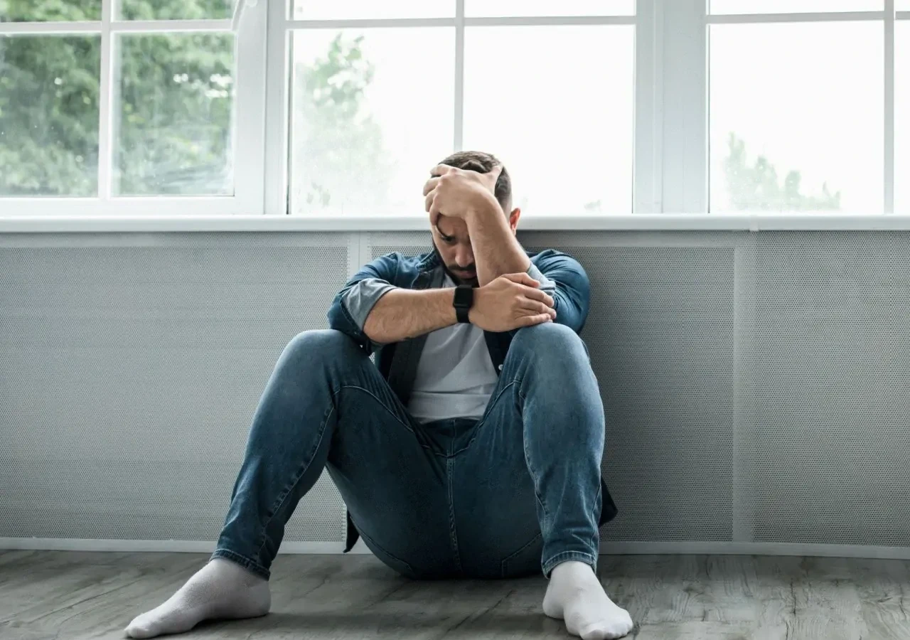 Depressieve man met spijkerbroek en witte sokken zittend tegen een raam met hand op het hoofd. 
