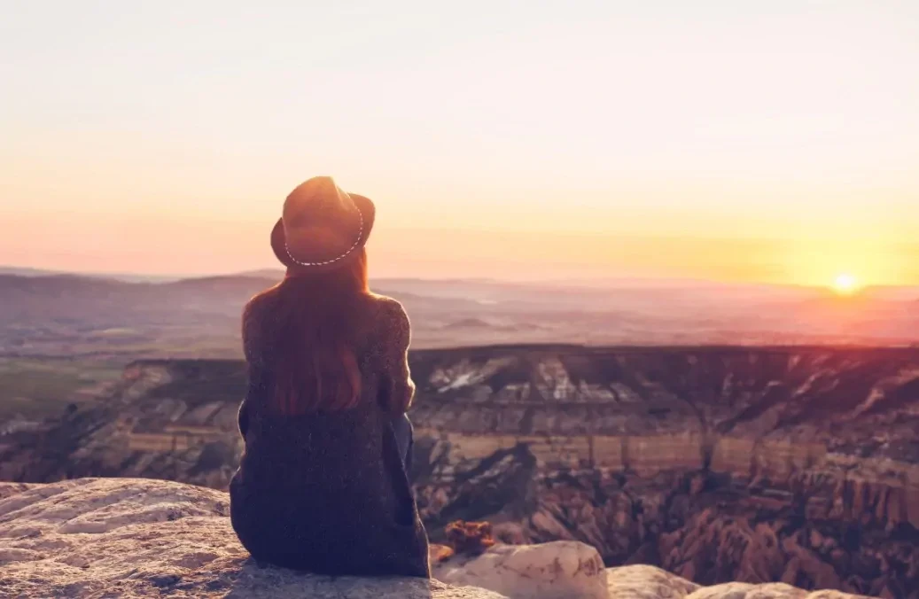 Vrouw zit op een rots en kijkt naar de zonsondergang over een natuurgebied uit.