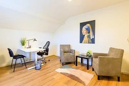 Behandelkamer met twee fauteuils, een salontafeltje, een bureau en twee bureaustoelen met op de achtergrond een schilderij.