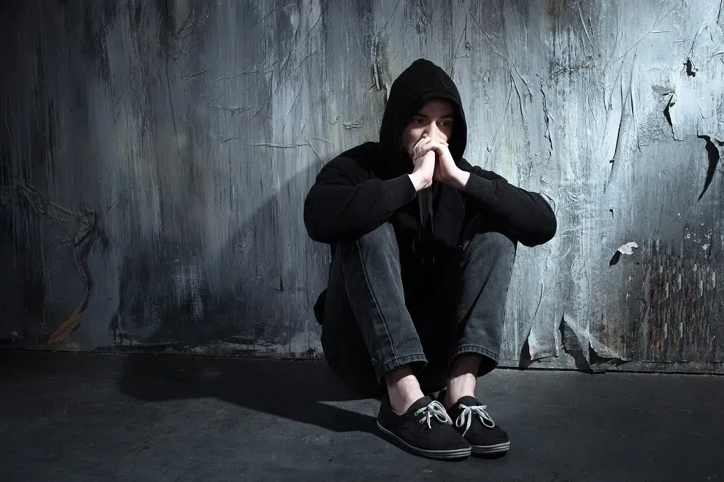 Depressieve man zittend tegen een verweerde muur in donkere kleding met capuchon op en handen half voor het gezicht.