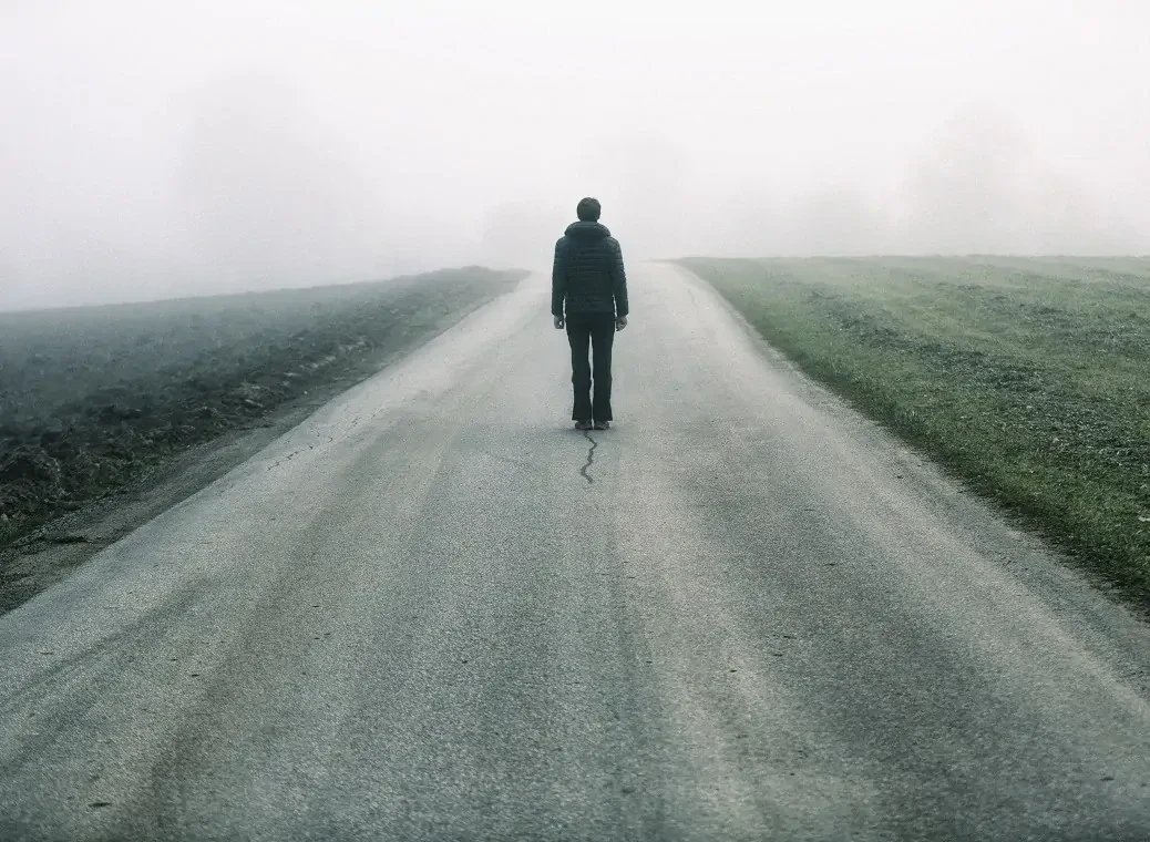 Eenzame man lopend met zijn rug naar de kijken op een lege weg, met daarnaast grasvelden in een grauwe omgeving. 