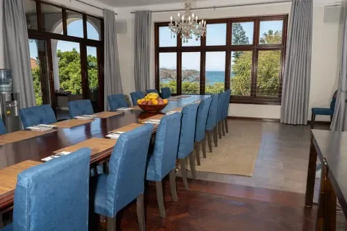 Vergaderruimte met lange houten tafel met daaraan blauwe stoelen en uitzicht via verschillende ramen op een groene omgeving en de zee.