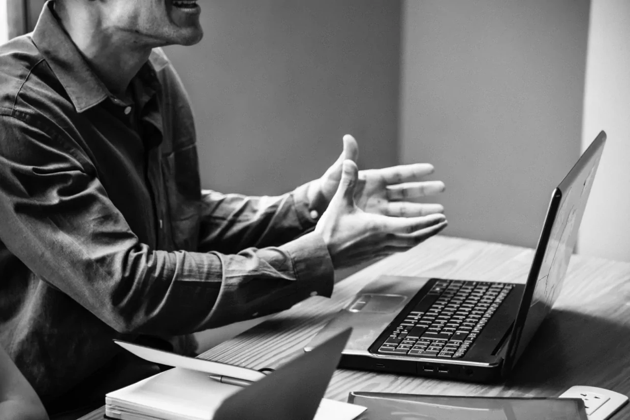 Zwart-wit foto van een man achter een bureau die de handen vragend uitstrekt en achter een laptop zit.