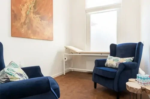 Behandelkamer met witte muren en een schilderij aan de muur, twee donkerblauwe fauteuils en een houten bijzettafeltje.