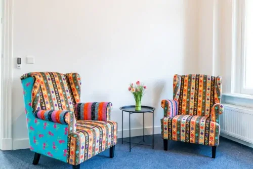 Behandelruimte met witte muren, een blauw tapijt en twee opvallend bedrukte fauteuils met daartussen een bijzettafeltje.