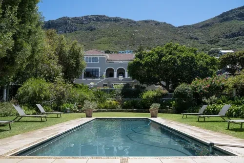 Tuin met ligbedden op het gras aan weerskanten van het zwembad, met op de achtergrond een villa, bergen en een strak blauwe lucht.