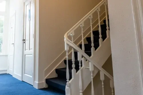 Gang met blauw tapijt, witte muren en een trap die naar boven leidt.
