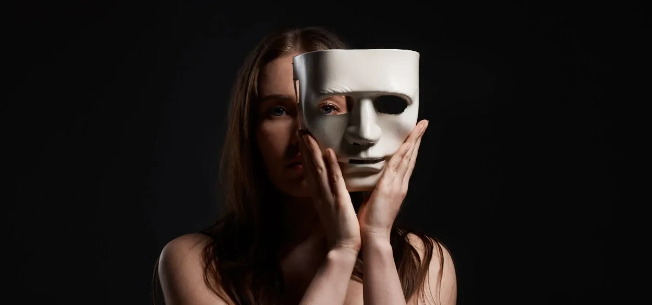 Vrouw met bruin haar houdt in twee handen een wit masker half voor haar gezicht. 