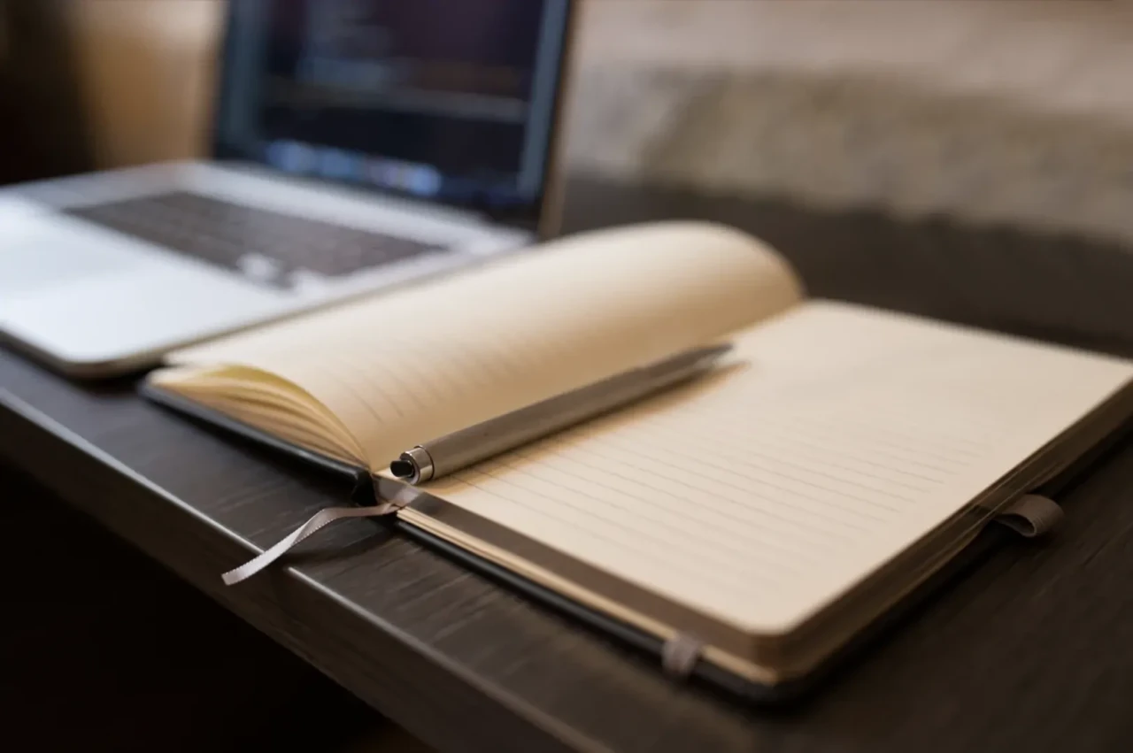 Notitieboekje ligt open op een bureau met een pen erbij en een laptop ernaast.