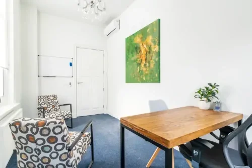 Behandelruimte met witte muren en een groen schilderij, een blauw tapijt, een houten bureau en twee stoelen in een drukke print.