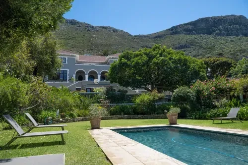 Tuin met ligbedden in het gras naast een zwembad, met op de achtergrond een villa, bergen en strak blauwe lucht.