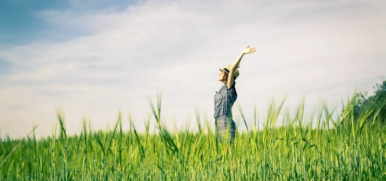 Vrouw met strohoed met de handen in de lucht in een groen grasveld en wolken en blauwe lucht op de achtergrond.