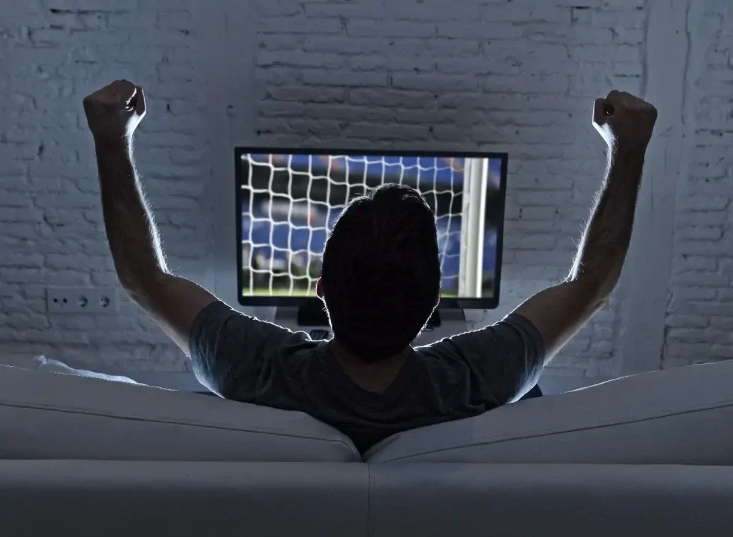Man op witte bank juicht nadat hij heeft gegokt op een voetbalwedstrijd, met tv op de achtergrond.