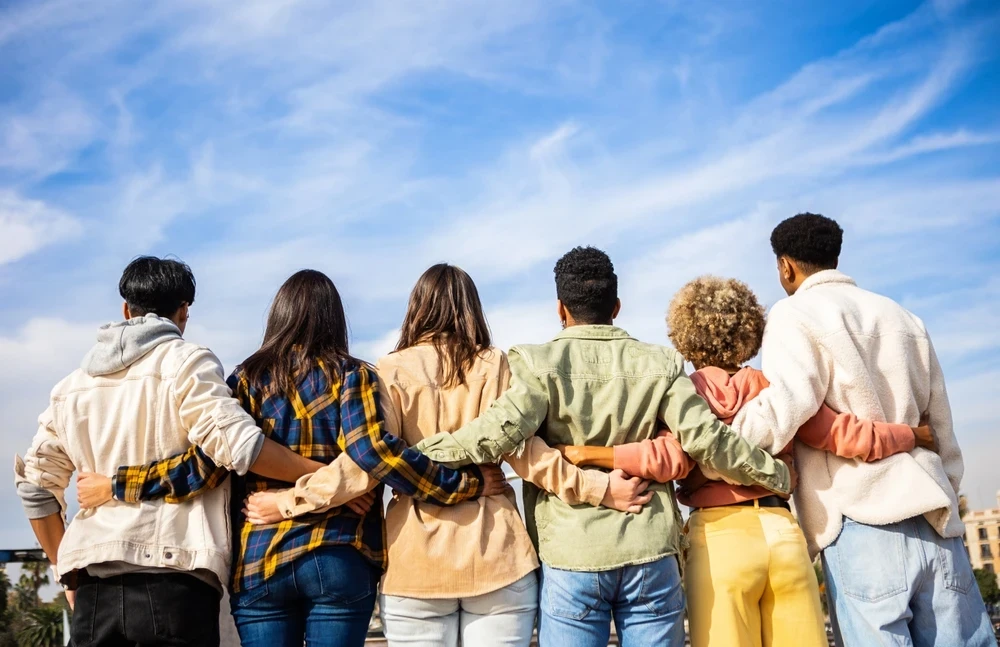 Een groep van zes mensen op een rij die elkaar vasthouden en van de camera afkijken naar een blauwe lucht.