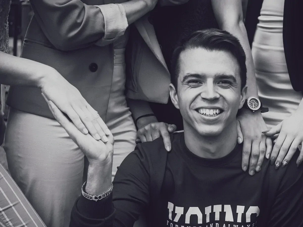 Zwart-wit foto van blije jongen die omringd wordt door mensen die hun handen op zijn schouders leggen.