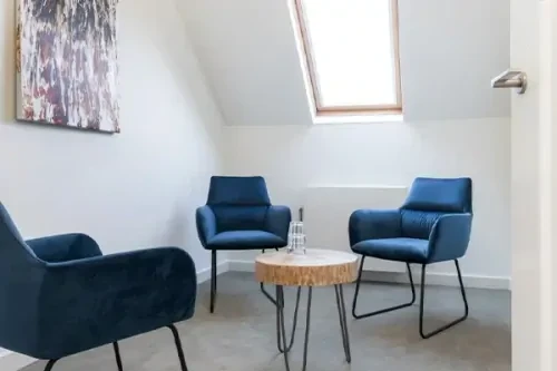 Behandelkamer met drie donkerblauwe stoelen met daartussen een bijzettafeltje, witte muren en een schilderij.