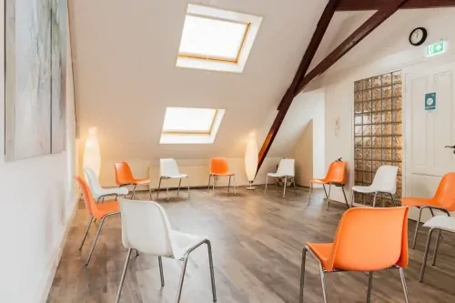 Behandelkamer voor kringgesprekken met witte en oranje stoelen in kringopstelling op een houten vloer en met witte muren.