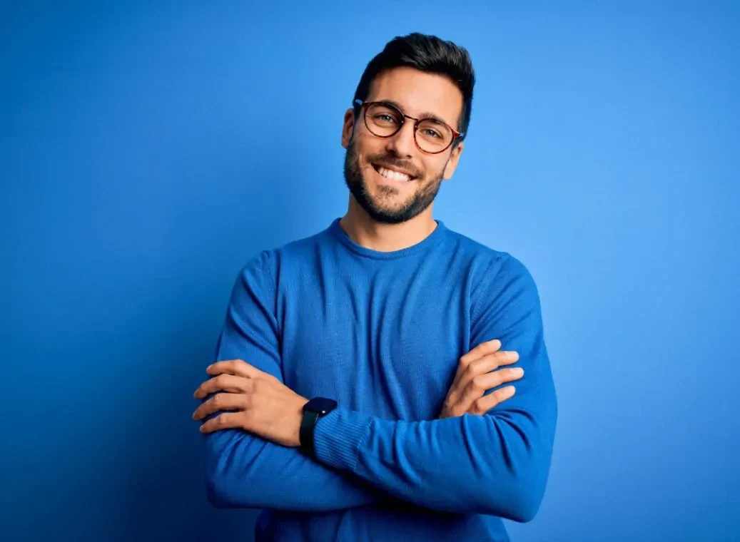 Lachende man met bril op en een blauwe trui aan staat voor een blauwe achtergrond met de armen over elkaar.