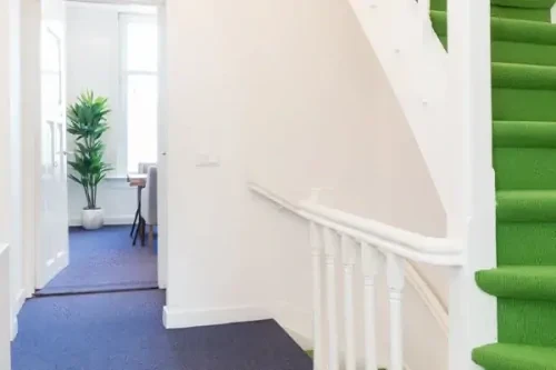 Hal met witte muren, een blauw tapijt en een groene trap die naar boven leidt.