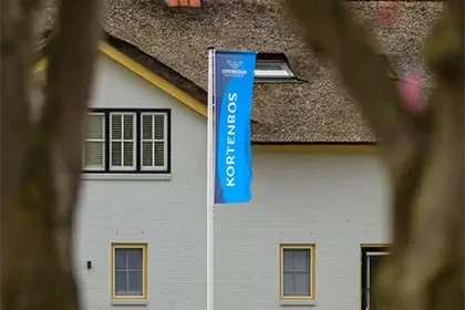 Blauwe vlag met daarop "kortenbos' geschreven aan een witte paal, voor een wit huis met rieten dak en bomen uit focus op de voorgrond.